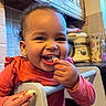 appliance, cabinet, child, curly_hair, cute, eating, face, food, furniture, hand, happy, high_chair, indoors, kitchen, person, smiling, snack, toddler, warm_lighting, young_child