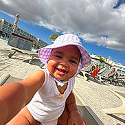 Ophelia joined the competition — help win amazing prizes! baby, child, hat, purple_hat, white_clothing, smiling, outdoor, poolside, lounge_chair, sky, clouds, resort, sunlight, happy, toddler, bracelet, vacation, person, summer, playful