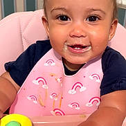 Felicity joined the competition — help win amazing prizes! baby, bib, child, curly_hair, cute, face, food_stains, green_buttons, hand, happy, high_chair, indoor, infant, messy_face, pink_chair, portrait, rainbow_pattern, smiling, toy, yellow_toy