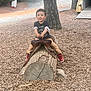 Carlos is registered to the contest to win money with this photo: child, boy, wood_log, saddle, playground, wood_chips, tree, outdoor, nature, casual_clothing, red_shoes, smiling, pretend_play, daytime, park, recreation, young_child, fun, adventure, woodland