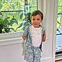 child, boy, indoor, window, plants, greenery, white_wall, wooden_floor, shorts, shirt, tshirt, necklace, smiling, standing, natural_light, casual_clothing, home, background, happy, portrait