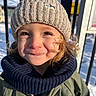 child, winter_clothing, knit_hat, scarf, smile, face, outdoor, snow, sunlight, curly_hair, jacket, portrait, happy, cold_weather, daylight, person, headwear, young_child, blue_sky, fence