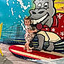 Hannah joined the competition — help win amazing prizes! toddler, child, smiling, surfboard, mural, donkey, cartoon, wall_art, indoor, flooring, yellow_chair, colorful, playful, dress, footwear, hair_bow, painting, person, happy, art