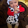 baby, baby_rocker, blanket, bow, brown_blanket, child, couch, cozy, face, hands, happy, headband, indoor, infant, polka_dot_dress, portrait, red_sweater, sitting, smiling, toy