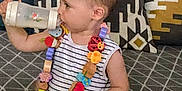Noam a rejoint le concours — aidez-le/la à gagner de superbes lots ! toddler, child, bottle, drinking, striped_shirt, necklace, wooden_beads, colorful, couch, pillows, patterned, geometric, blanket, indoor, person, young_child, furniture, home, casual, cute