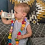 toddler, child, bottle, drinking, striped_shirt, necklace, wooden_beads, colorful, couch, pillows, patterned, geometric, blanket, indoor, person, young_child, furniture, home, casual, cute