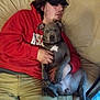 person, dog, couch, hoodie, cap, indoor, relaxed, pet, animal, man, casual, seated, living_room, gray_dog, white_patch, holding, resting, comfort, nap, bonding
