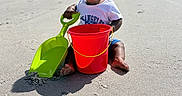 Kayson joined the competition — help win amazing prizes! toddler, beach, sand, bucket, shovel, pacifier, child, sunny_day, outdoor, play, toy, casual_clothing, happy, sitting, footprints, urban_background, sky, building, vacation, summer