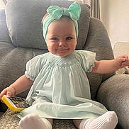 Nola is registered to the contest to win money with this photo: baby, child, smiling, headband, dress, armchair, toy, phone, socks, indoors, window, curtains, light, happy, cute, infant, seated, person, furniture, cozy