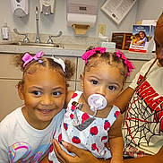 Saige is registered to the contest to win money with this photo: child, baby, toddler, pacifier, hair_bows, smiling, medical_office, sink, counter, hand_sanitizer, brother_or_sister, person, indoor, clothing, cute, holding, family, young_girl, healthcare, infant