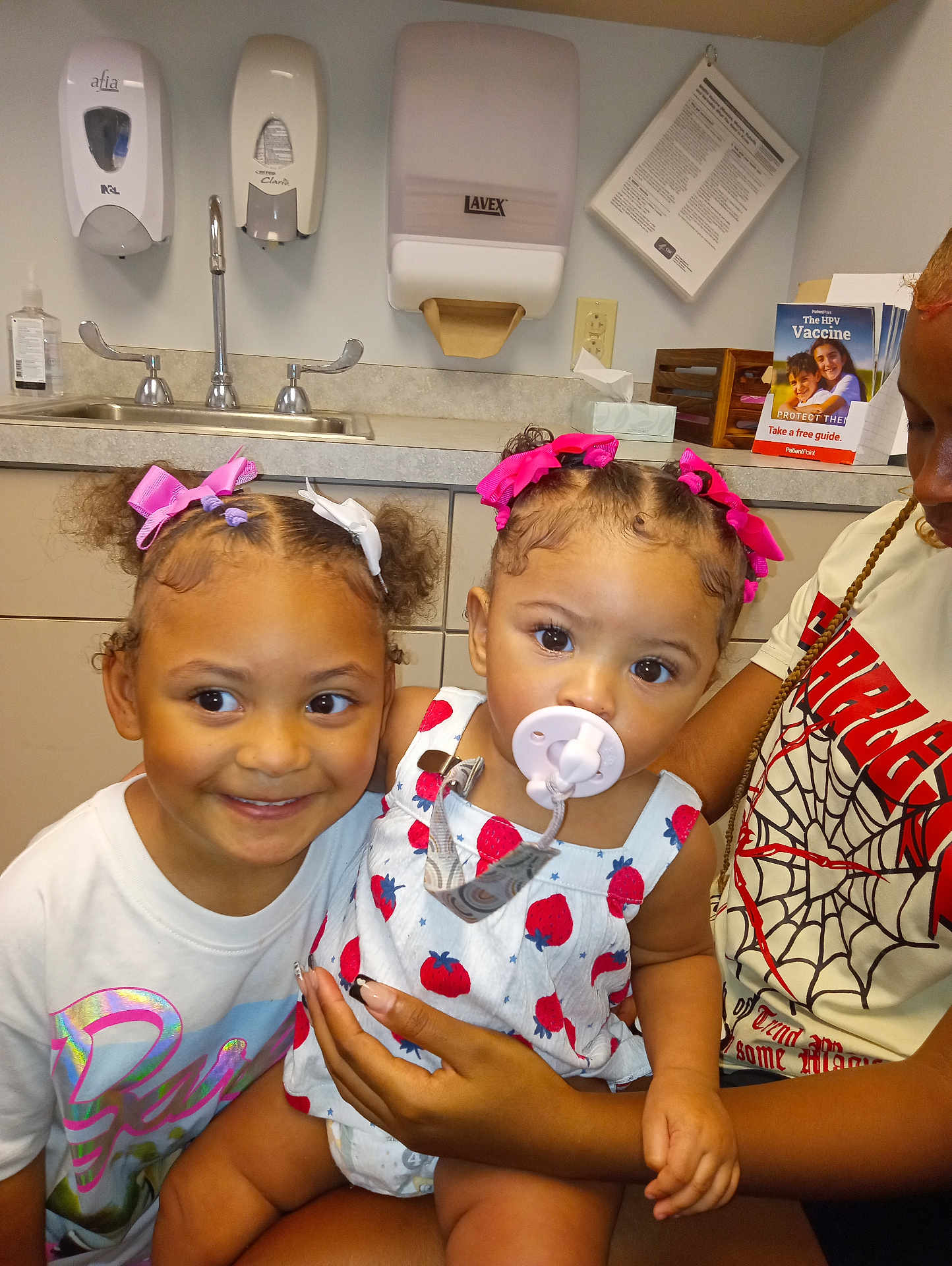 Saige is registered to the contest to win money with this photo: child, baby, toddler, pacifier, hair_bows, smiling, medical_office, sink, counter, hand_sanitizer, brother_or_sister, person, indoor, clothing, cute, holding, family, young_girl, healthcare, infant