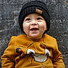 baby, child, smiling, sitting, black_hat, yellow_sweatshirt, cross_necklace, concrete_wall, outdoor, footwear, happy, portrait, cute, clothing, person, infant, casual, urban, expression, young