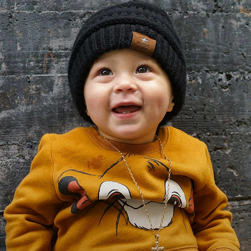 Tauren joined the competition — help win amazing prizes! baby, child, smiling, sitting, black_hat, yellow_sweatshirt, cross_necklace, concrete_wall, outdoor, footwear, happy, portrait, cute, clothing, person, infant, casual, urban, expression, young