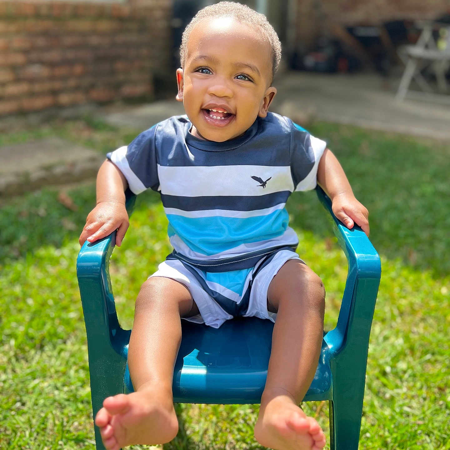 Brexton is registered to the contest to win money with this photo: toddler, child, smiling, blue_chair, outdoor, grass, sunlight, happy, shorts, striped_shirt, barefoot, plastic_chair, daylight, casual_clothing, portrait, young_child, sitting, greenery, cheerful, playful