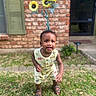 baby, brick_wall, child, curious_expression, daylight, door, garden, grass, greenery, outdoor, person, playful, romper, sandals, standing, summer, sunflower_art, toddler, window, yellow_clothing