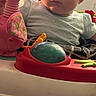 Deliah is registered to the contest to win money with this photo: baby, child, toy, dinosaur_toy, baby_walker, blue_bow, sneakers, carpet, indoor, person, cute, infant, playtime, colorful, young_child, clothing, footwear, expression, holding, furniture