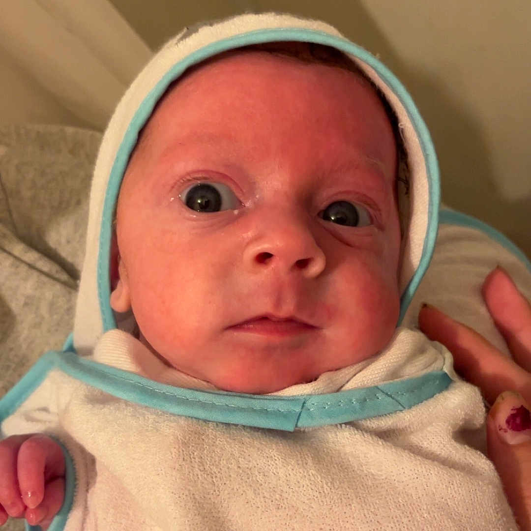 Halton is registered to the contest to win money with this photo: baby, blue_trim, closeup, clothing, curious, expression, eyes, face, hand, headwear, indoors, infant, newborn, person, portrait, skin, soft_texture, support, towel, wrapped