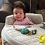 baby, infant, child, spiky_hair, tongue_out, baby_seat, play_mat, couch, cushion, living_room, cozy, patterned, soft_furniture, pink_clothing, cute, happy, portrait, indoor, person, smiling