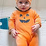 baby, pumpkin_costume, orange_clothing, infant, cute, play_mat, face, hat, footed_pajamas, child, looking, lying_down, indoors, patterned_background, chubby_cheeks, wide_eyes, costume, holiday, smiling, one_person