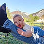 baby, child, person, legs, jeans, socks, outdoor, grass, mountain, sky, tree, house, bench, daylight, nature, playful, expression, casual_clothing, greenery, happy