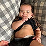 baby, child, smiling, shirt, diaper, hand, cushion, checkered, black_clothing, skin, face, lying_down, cheeks, comfort, indoor, cute, infant, playful, relaxing, person