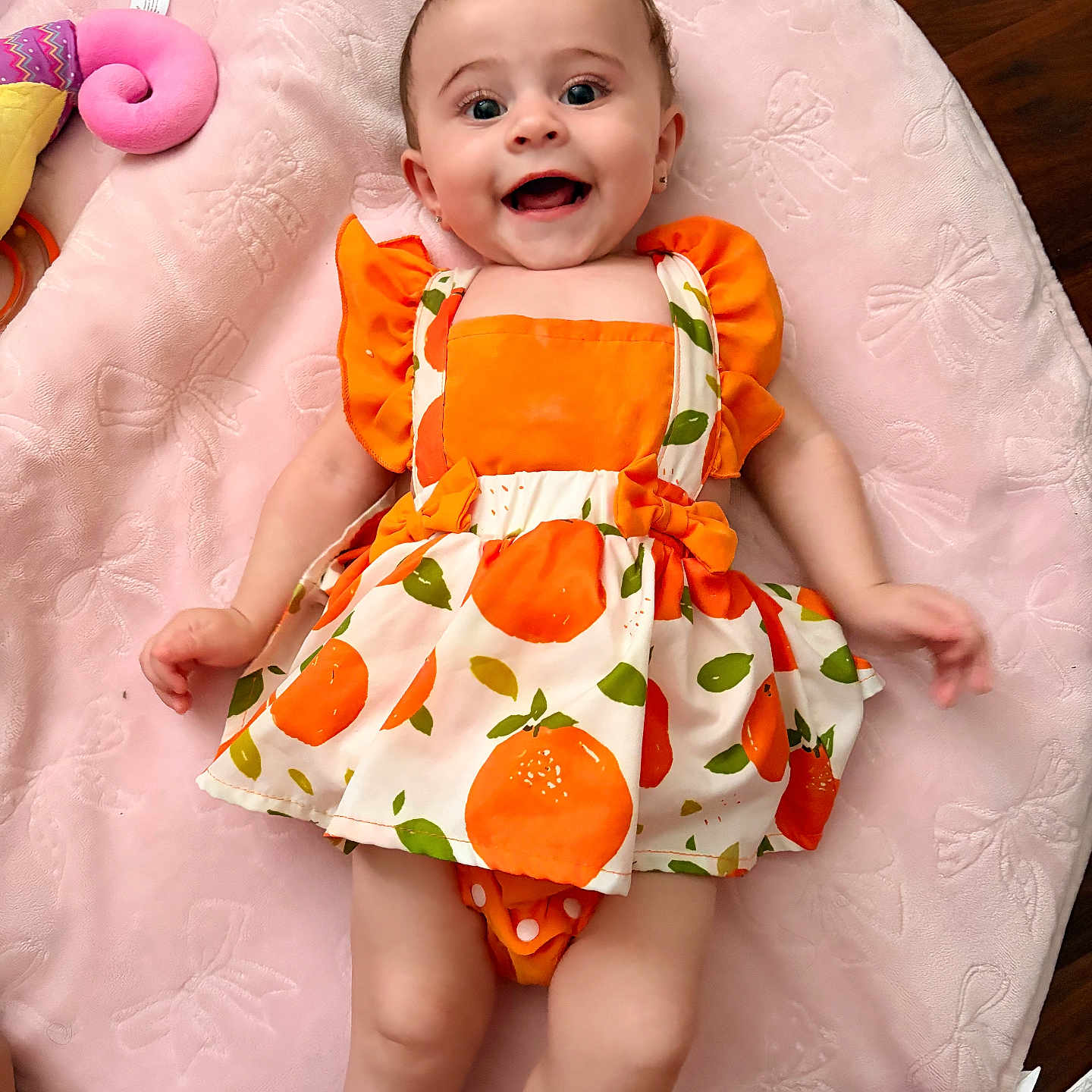 Solena is registered to the contest to win money with this photo: arms, baby, child, cute, dress, eyes, face, floor, happy, indoors, infant, laying_down, legs, orange, patterned_clothing, pink_blanket, playful, plush_toy, smile, soft_texture