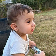 Said is registered to the contest to win money with this photo: toddler, child, baby, portrait, profile, outdoor, garden, backyard, grass, trees, fence, chair, sitting, curious, side_view, short_hair, white_shirt, snack, pouch, daylight
