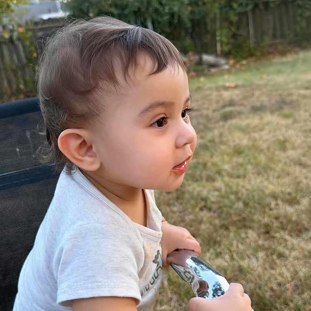 Said is registered to the contest to win money with this photo: toddler, child, baby, portrait, profile, outdoor, garden, backyard, grass, trees, fence, chair, sitting, curious, side_view, short_hair, white_shirt, snack, pouch, daylight