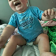 Ridley is registered to the contest to win money with this photo: baby, child, smiling, blue_shirt, diaper, bed, blanket, indoor, happy, reaching, skin, face, person, young_child, cute, expression, infant, sitting, comfort, clothing