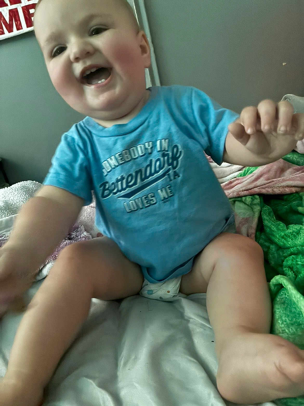 Ridley is registered to the contest to win money with this photo: baby, child, smiling, blue_shirt, diaper, bed, blanket, indoor, happy, reaching, skin, face, person, young_child, cute, expression, infant, sitting, comfort, clothing