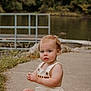 toddler, child, baby, outdoor, lake, water, concrete_path, grass, trees, greenery, romper, sitting, barefoot, curious, nature, summer, portrait, young_child, daylight, casual_clothing