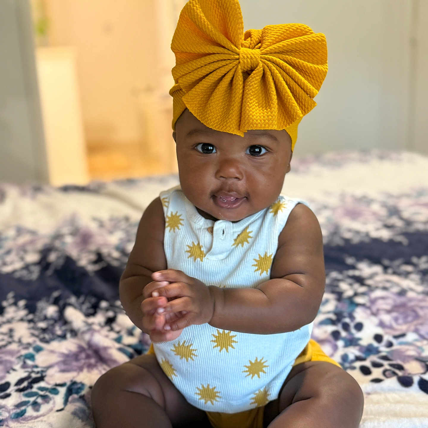 Dallis is registered to the contest to win money with this photo: apparel, baby, bedspread, child, clapping, cute, floral_pattern, furniture, happy, headband, indoors, infant, person, portrait, room, sitting, smiling, sparkly_shoes, sun_pattern, yellow