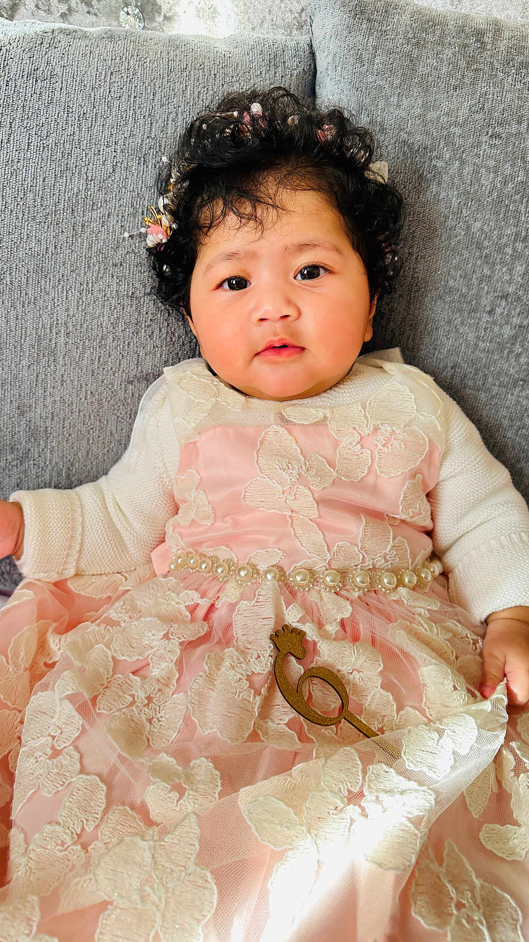 Devira is registered to the contest to win money with this photo: baby, curly_hair, flower_crown, pink_dress, lace, pearl_belt, couch, gray_background, infant, portrait, cute, indoors, child, soft_lighting, fashion, hand, expression, sitting, closeup, person