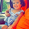 child, toddler, smiling, portrait, car, vehicle, passenger, headphones, dress, floral_pattern, sitting, indoor, travel, family, adult, person, window, daylight, orange_shirt, happy
