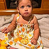 child, toddler, girl, baby, portrait, sitting, dress, floral_pattern, fruit, orange, lemon, citrus, bed, indoors, home, face, eyes, braids, earrings, photography