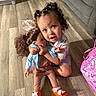 child, toddler, girl, doll, toy, hugging, sitting, smiling, curly_hair, hair_bows, indoor, living_room, wood_floor, sofa, backpack, colorful_clothing, orange_shoes, portrait, playtime, family