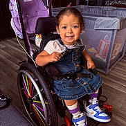Mazikeen is registered to the contest to win money with this photo: child, colorful, denim_dress, family, girl, happy, home, indoors, playpen, playroom, portrait, rainbow, seated, smiling, sneakers, striped_shirt, stroller, toddler, wheel, wheelchair