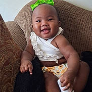 Jilisha joined the competition — help win amazing prizes! baby, child, smiling, green_bow, white_top, floral_bottoms, socks, brown_chair, happy, cute, indoors, infant, portrait, person, seated, cozy, comfortable, tongue_out, playful, young