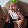 baby, child, smiling, green_bow, white_top, floral_bottoms, socks, brown_chair, happy, cute, indoors, infant, portrait, person, seated, cozy, comfortable, tongue_out, playful, young