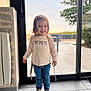 child, girl, smiling, standing, indoors, glass_door, jeans, sneakers, hair_clip, sweater, floor_tiles, window, natural_light, backpack, happy, casual_clothing, portrait, young_child, home_interior, daylight