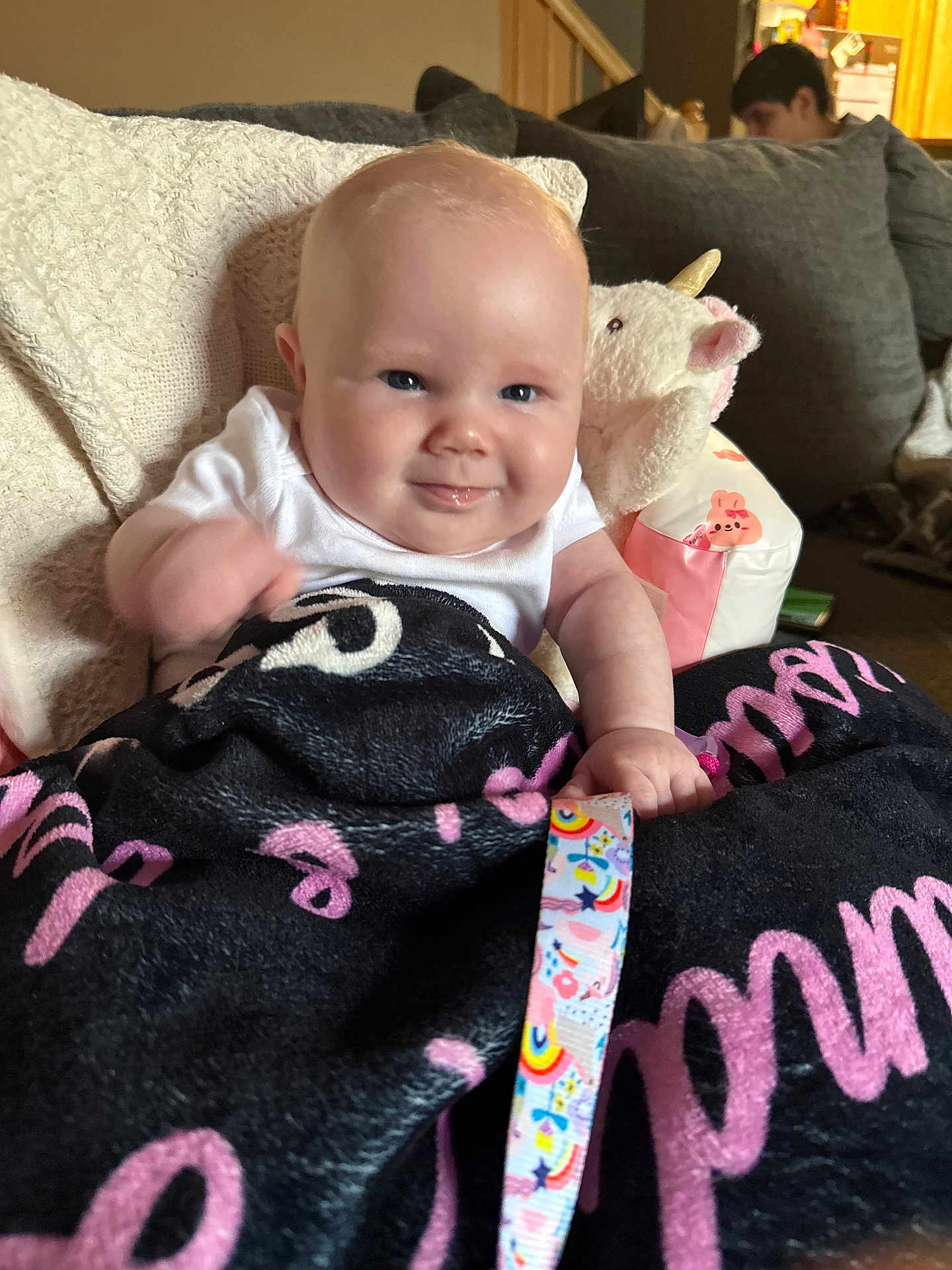 Isabella is registered to the contest to win money with this photo: baby, smile, blanket, unicorn, plush_toy, couch, person, indoors, child, face, hand, expression, cozy, clothing, colorful, ribbon, furniture, home, cute, portrait