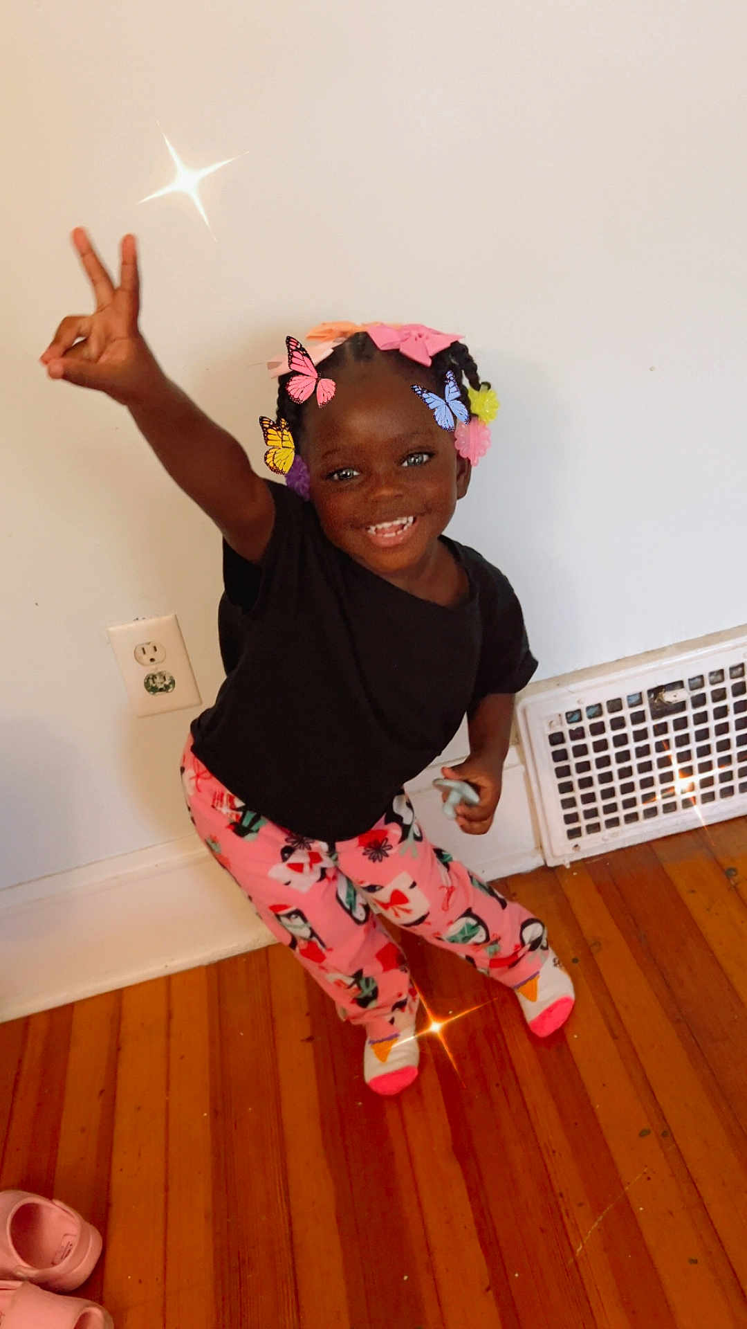 Ava joined the competition — help win amazing prizes! child, smiling, peace_sign, indoor, colorful, butterflies, hair_accessories, black_tshirt, pink_pants, patterned_clothing, wooden_floor, white_wall, vent, socks, happy, standing, playful, toy, footwear, young_kid