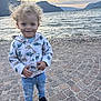 child, clouds, cobblestone, curly_hair, dinosaurs, happy, hoodie, jeans, lake, mountains, nature, outdoor, person, portrait, sky, smiling, sneakers, sunset, toddler, water