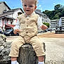 beige_clothing, bow_tie, buildings, cars, child, curly_hair, daytime, happy, outdoor, pants, portrait, rock, sky, smiling, toddler, trees, urban, vest, white_shoes, white_socks
