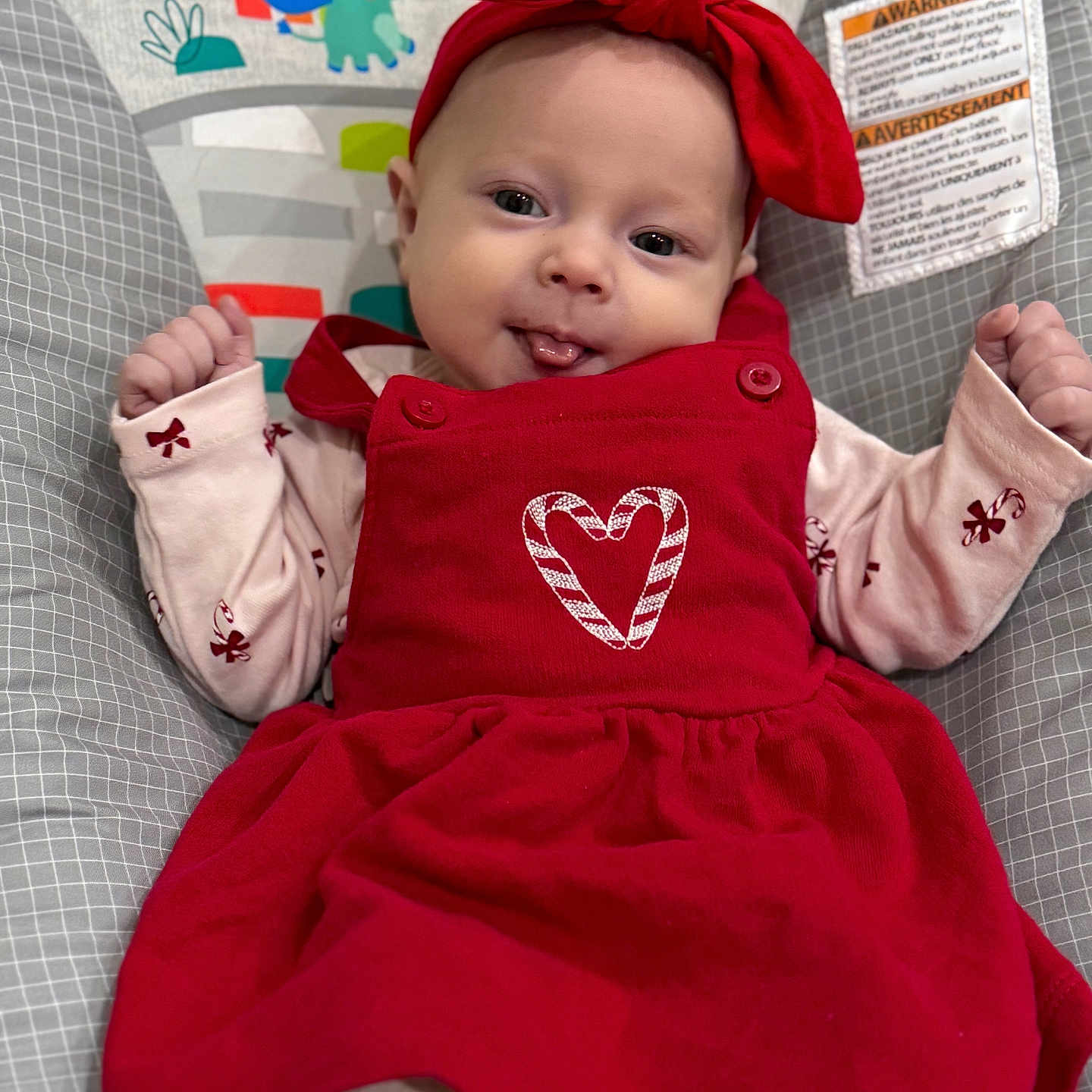Hazel is registered to the contest to win money with this photo: baby, baby_seat, cartoon_animals, child, clothing, colorful, cute, fabric, happy, headband, indoors, infant, pattern, person, playful, portrait, red_dress, smiling, tongue_out, young