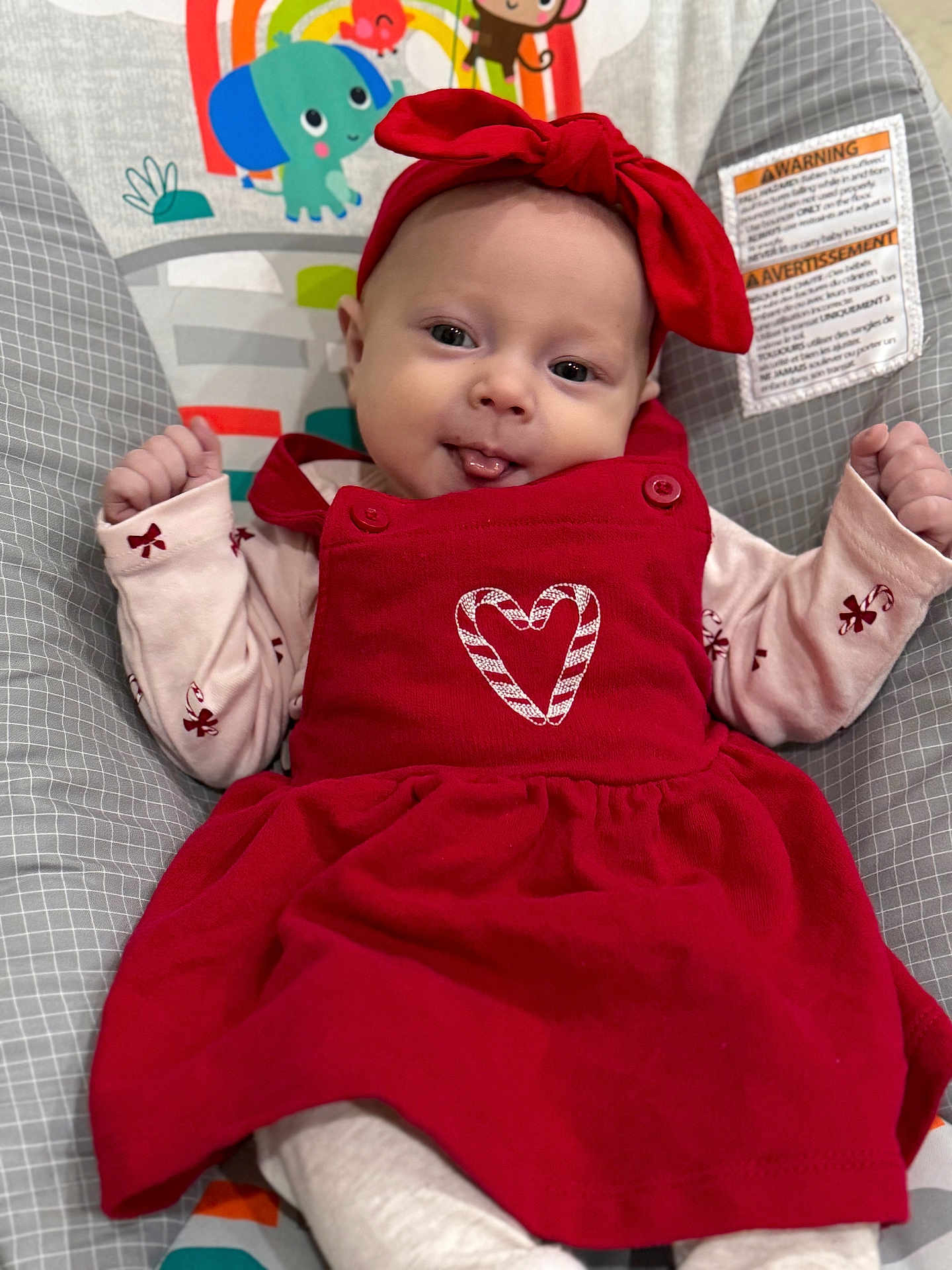 Hazel is registered to the contest to win money with this photo: baby, infant, red_dress, headband, tongue_out, baby_seat, cartoon_animals, cute, playful, clothing, pattern, smiling, child, indoors, colorful, person, fabric, happy, portrait, young