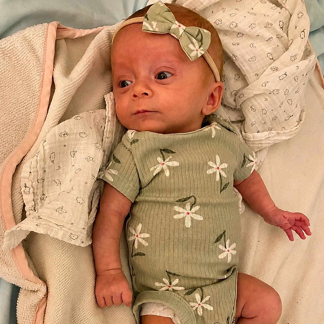 Allie is registered to the contest to win money with this photo: baby, infant, child, green_onesie, headband, bow, blanket, soft_texture, cute, newborn, lying_down, clothing, patterned, floral, sleepwear, indoors, skin, hand, face, portrait