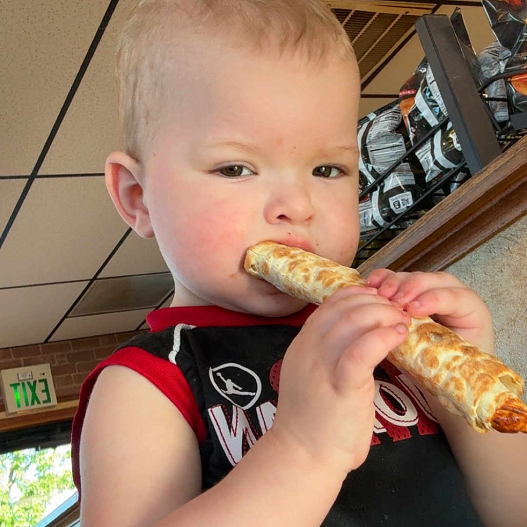 Geovanni is registered to the contest to win money with this photo: black_shirt, breadstick, ceiling, child, clothing, daylight, eating, exit_sign, face, food, hands, indoor, red_trim, restaurant, serious_expression, short_hair, snacks, table, toddler, window
