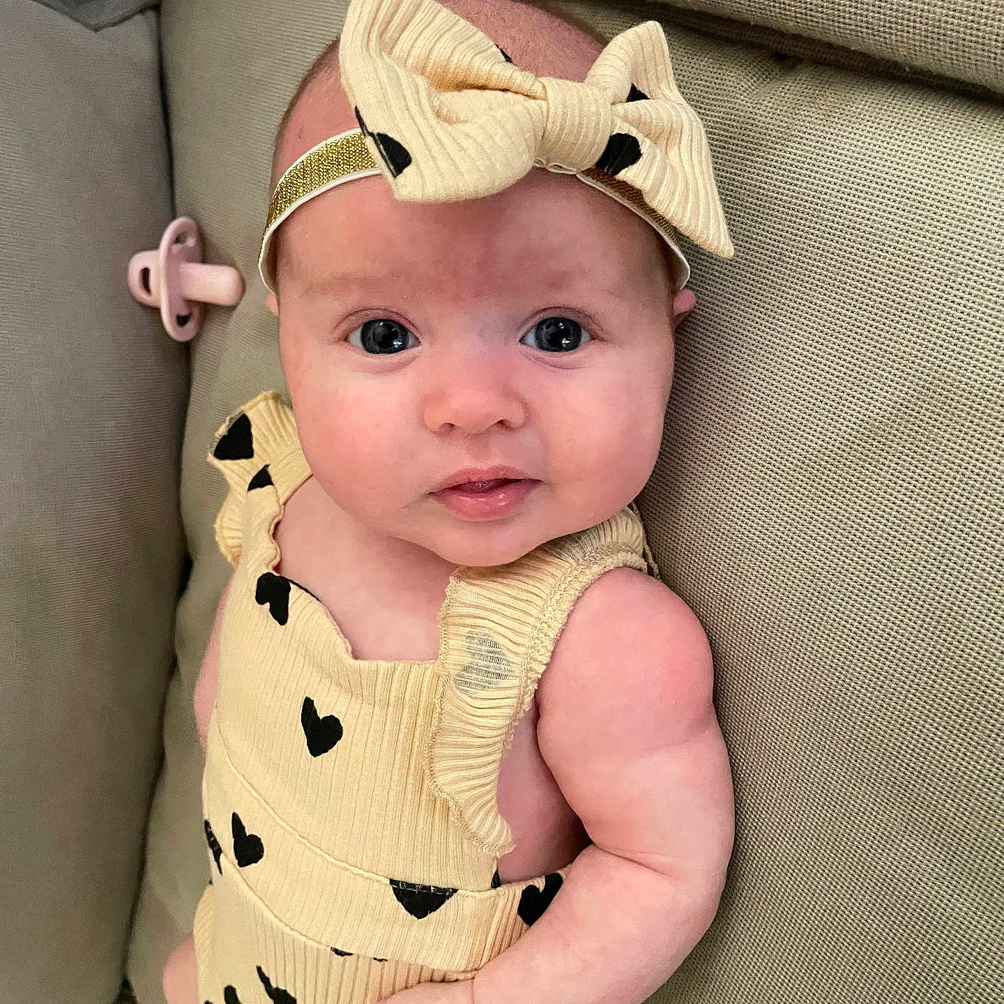Romey is registered to the contest to win money with this photo: baby, infant, child, headband, bow, yellow_outfit, black_hearts, couch, pacifier, skin, face, eyes, arm, furniture, comfortable, lying_down, curious, indoors, closeup, portrait