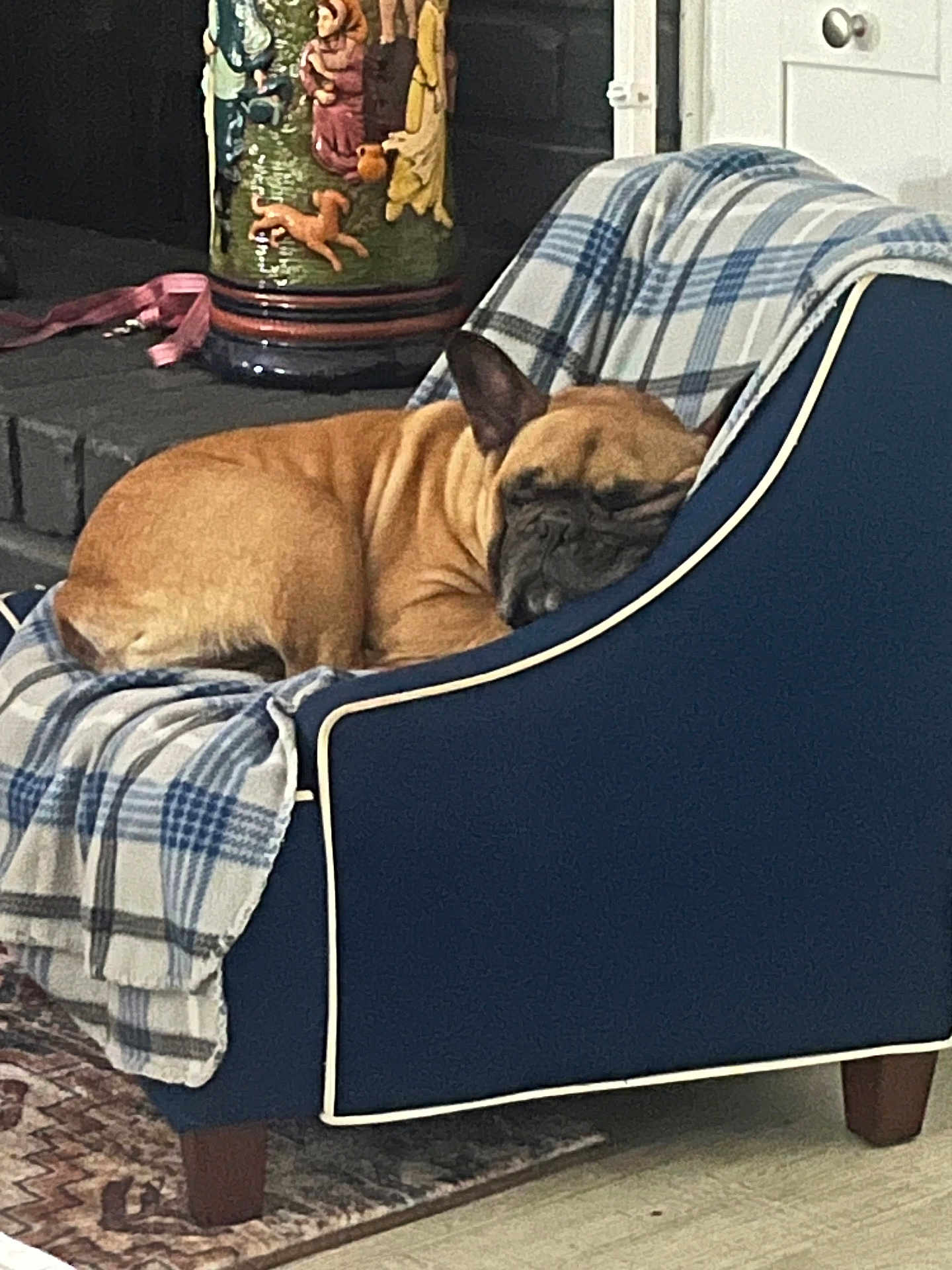 Puma is registered to the contest to win money with this photo: dog, french_bulldog, sleeping, armchair, plaid_blanket, indoor, vase, fireplace, decor, floor_rug, cozy, pet, resting, furniture, brown_dog, blue_chair, blanket, home, relaxing, napping